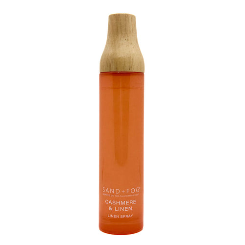 A tall cylindrical spray bottle of Sand + Fog Cashmere & Linen linen spray, featuring a translucent orange body and a natural light wood cap, set against a plain white background.