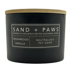 Black cedarwood vanilla candle with a wooden lid labeled 'Sand + Paws' on a white background
