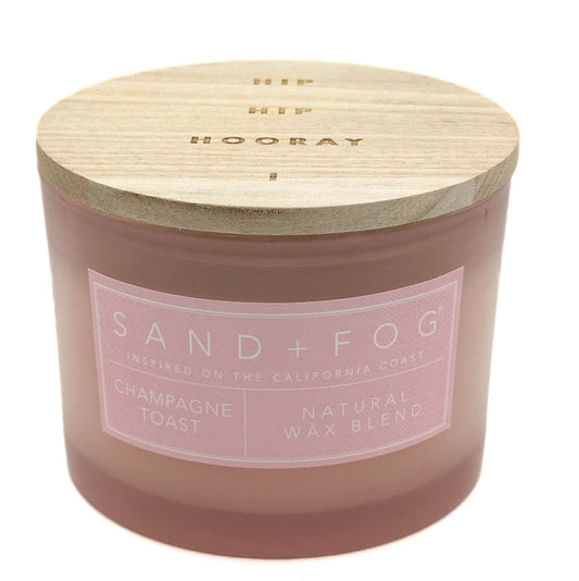 A Sand + Fog candle in a frosted glass jar with a light wood lid, featuring a pink label that reads 'Champagne Toast' and 'Natural Wax Blend'.