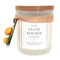 A 'Clean Kitchen' scented candle by DW Home in a frosted glass jar with a cork lid, wrapped in a rustic jute ribbon. The candle is decorated with two small oranges and green leaves on the left side, set against a plain white background.