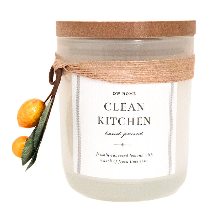 A 'Clean Kitchen' scented candle by DW Home in a frosted glass jar with a cork lid, wrapped in a rustic jute ribbon. The candle is decorated with two small oranges and green leaves on the left side, set against a plain white background.