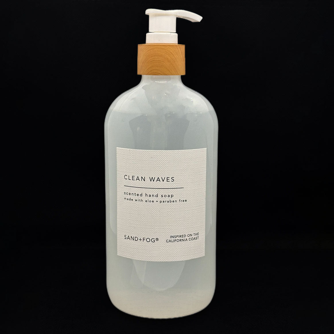 SAND + FOG Hand soap bottle labeled 'Clean Waves' with a pump and wooden cap on a black background