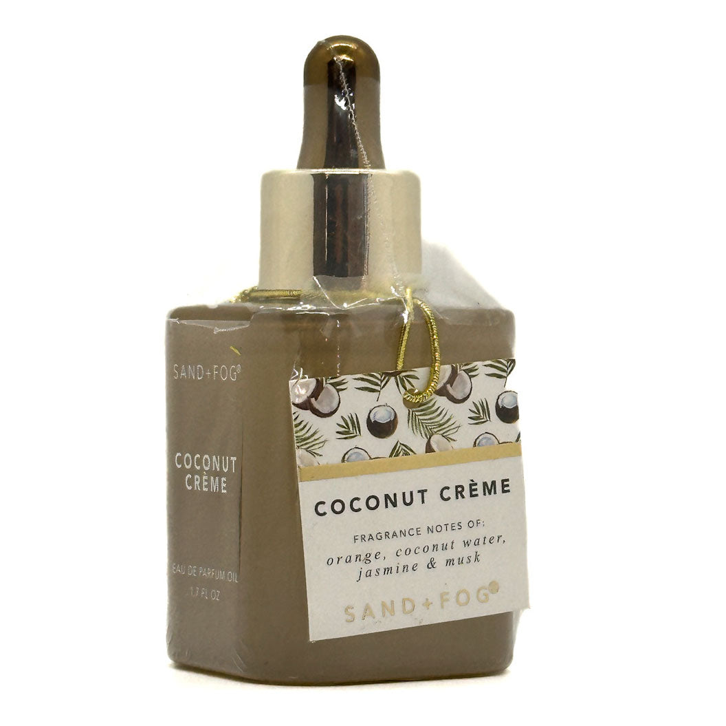 A bottle of Sand+Fog Coconut Crème fragrance oil with a brown body, gold dropper cap, and a decorative white and gold label, shown on a white background.