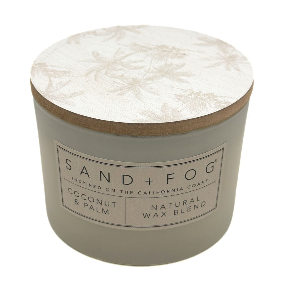 A round candle jar with a light beige body and a matching lid featuring a subtle palm tree pattern. The front label reads 'SAND + FOG' and 'COCONUT & PALM NATURAL WAX BLEND'.