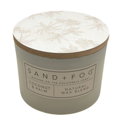 A round candle jar with a light beige body and a matching lid featuring a subtle palm tree pattern. The front label reads 'SAND + FOG' and 'COCONUT & PALM NATURAL WAX BLEND'.