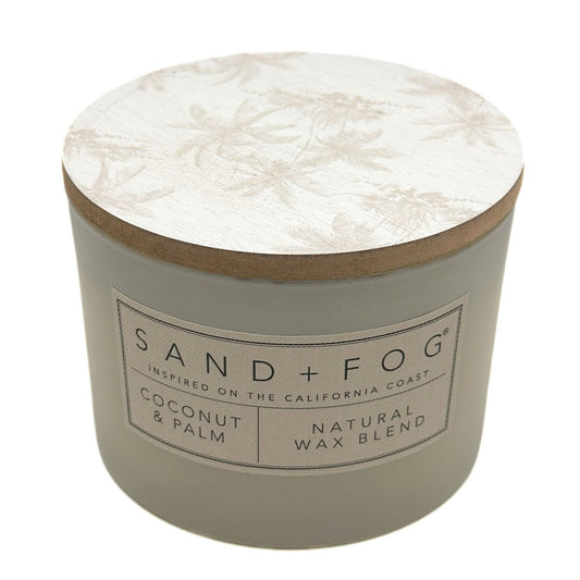 A round candle jar with a light beige body and a matching lid featuring a subtle palm tree pattern. The front label reads 'SAND + FOG' and 'COCONUT & PALM NATURAL WAX BLEND'.