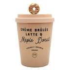A beige insulated coffee cup with a lid, featuring a donut-shaped metal charm on top. The cup is printed with the text 'CRÈME BRÚLÉE LATTE & Maple Donut' and a 'DW Design' logo, on a solid white background.