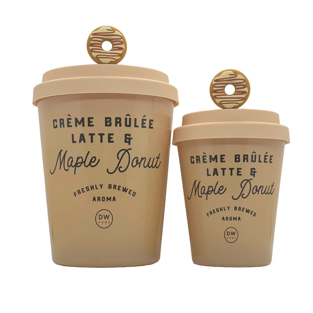 Two beige ceramic coffee cups shaped like latte cups, one large and one small, both with matching lids and round donut-shaped handles. The cups are printed with 'CRÈME BRÛlÉE LATTE & Maple Donut' and a small 'DW' logo, set against a plain white background.
