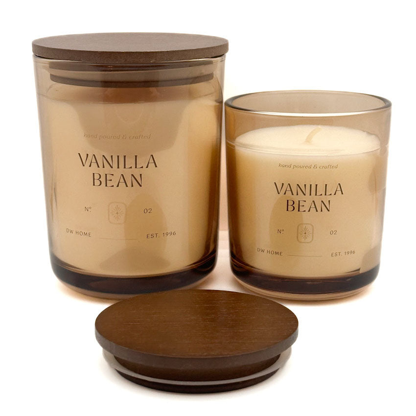 Two glass jar of DW Home candles labeled 'VANILLA BEAN' with warm cream-colored wax, shown on a white background. One larger jar has a dark brown wooden lid, and a smaller matching glass candle with its lid placed in front.