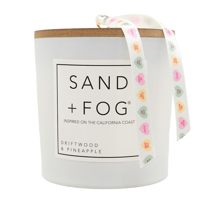 Driftwood & Pineapple candle with 'Sand + Fog' branding and a colourful ribbon on a white background