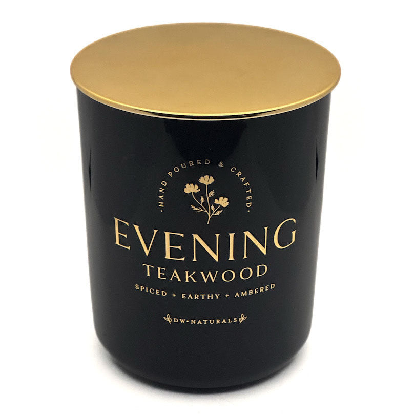 A dark green glass jar candle with a metallic gold lid, featuring gold lettering that reads 'EVENING TEAKWOOD' and 'SPICED + EARTHY + AMBERED' on a clean white background.