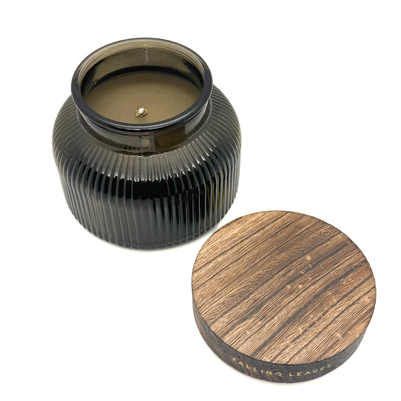 An open ribbed dark glass candle jar with a wooden lid placed beside it on a white background. The lid is engraved with the scent name 'FALLING LEAVES'.