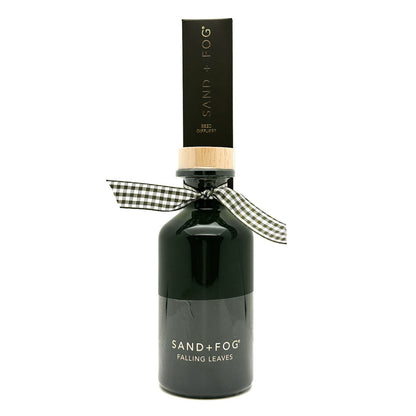A dark glass bottle of Sand + Fog Falling Leaves reed Diffuser with a black cap, a light wood collar, and a black and white gingham ribbon bow, on a white background.