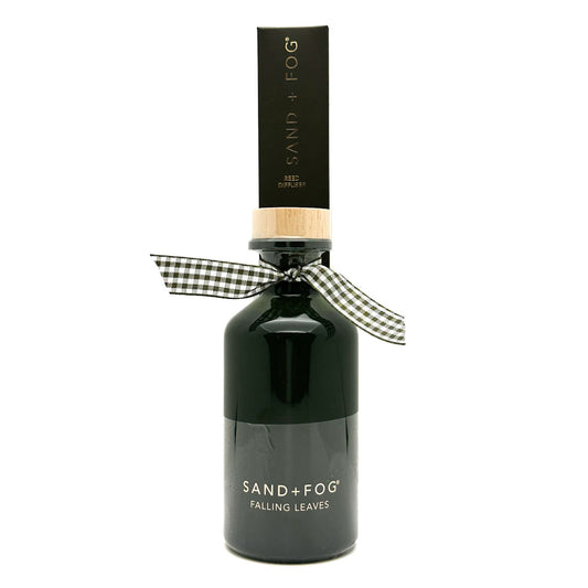 A dark glass bottle of Sand + Fog Falling Leaves reed Diffuser with a black cap, a light wood collar, and a black and white gingham ribbon bow, on a white background.