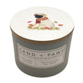 A cylindrical scented candle in a matte grey container with a round white lid printed with an illustration of a pug wearing a red scarf, surrounded by leaves. The front label reads 'SAND + PAWS' and 'FALLING LEAVES'.