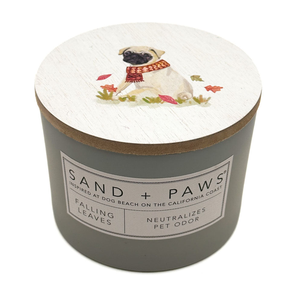 A cylindrical scented candle in a matte grey container with a round white lid printed with an illustration of a pug wearing a red scarf, surrounded by leaves. The front label reads 'SAND + PAWS' and 'FALLING LEAVES'.
