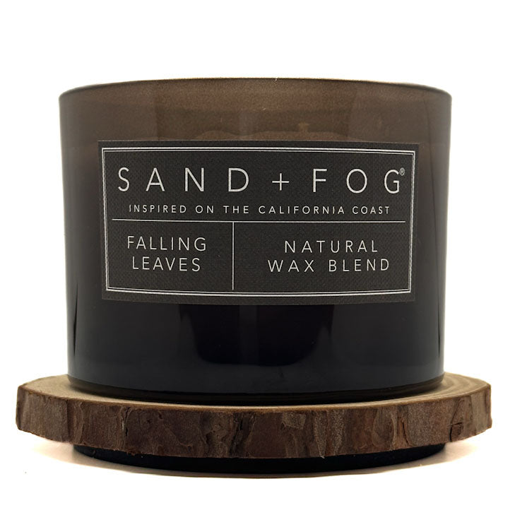 A dark brown translucent glass candle jar with a black label reading SAND+FOG, Falling Leaves, and Natural Wax Blend, resting on a round natural wood slice coaster against a white background.