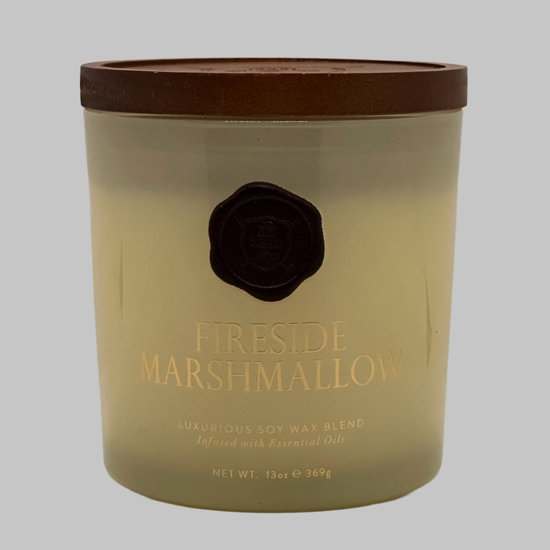A 13oz soy wax candle in a clear glass jar with a brown lid, featuring a gold foil label that reads 'FIRESIDE MARSHMALLOW' and a black wax seal, set against a solid grey background.