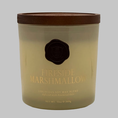 A 13oz soy wax candle in a clear glass jar with a brown lid, featuring a gold foil label that reads 'FIRESIDE MARSHMALLOW' and a black wax seal, set against a solid grey background.