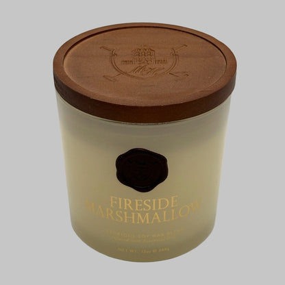 A 13oz soy wax candle in a clear glass jar with a brown lid with a crest on it, featuring a gold foil label that reads 'FIRESIDE MARSHMALLOW' and a black wax seal.