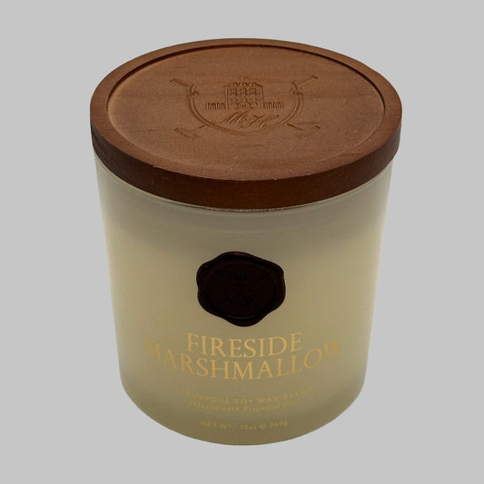 A 13oz soy wax candle in a clear glass jar with a brown lid with a crest on it, featuring a gold foil label that reads 'FIRESIDE MARSHMALLOW' and a black wax seal.