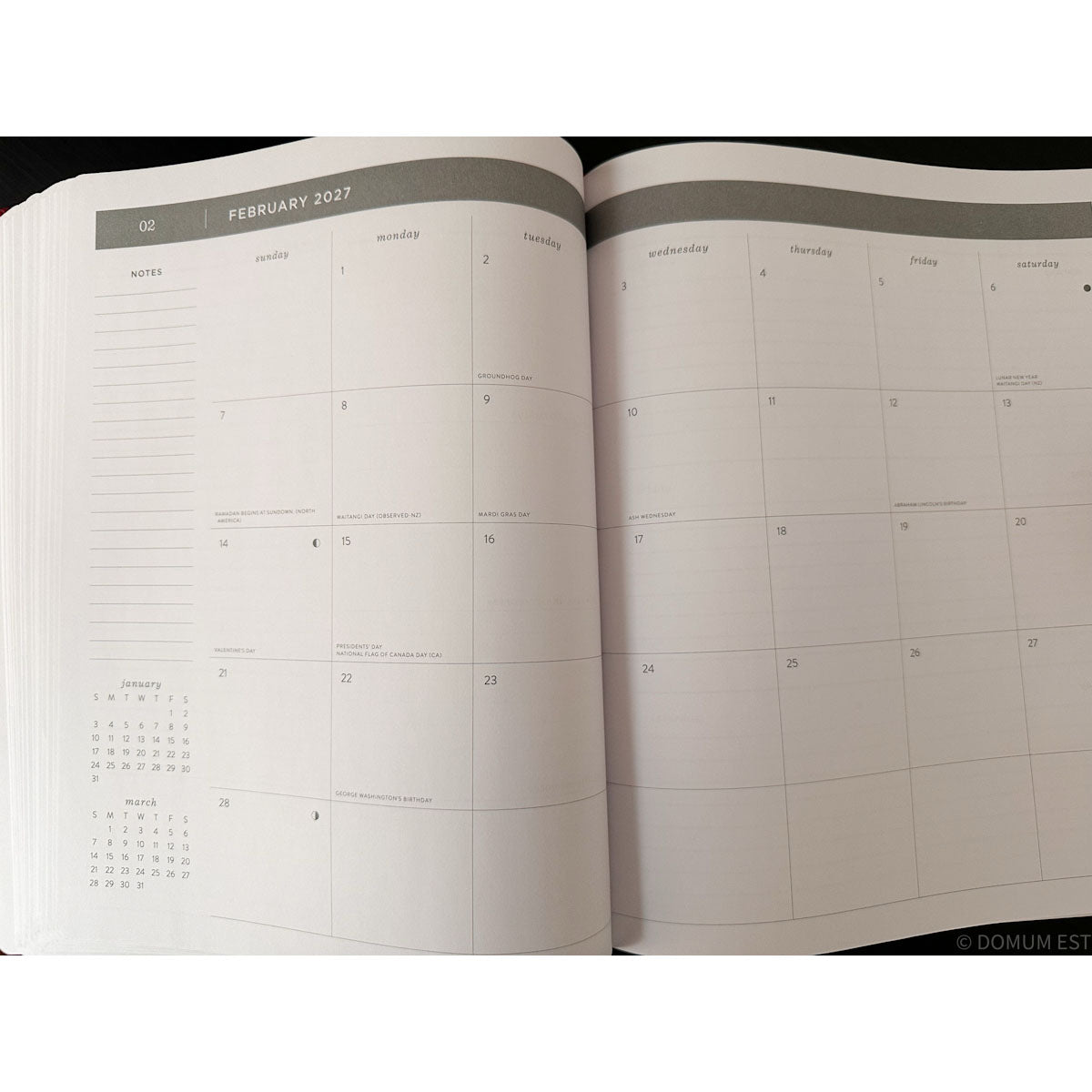 An open planner or calendar showing the February 2027 weekly layout with columns for Monday through Sunday and a small monthly calendar at the lower left, photographed against a dark background.