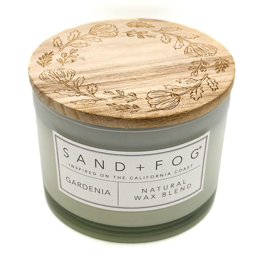 A nude glass jar candle with a natural bamboo lid engraved with botanical patterns. The front label reads 'SAND + FOG' and 'GARDENIA NATURAL WAX BLEND'.
