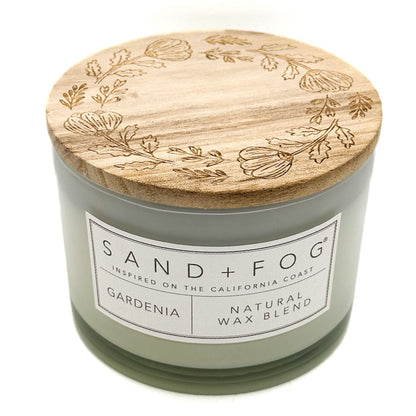 A nude glass jar candle with a natural bamboo lid engraved with botanical patterns. The front label reads 'SAND + FOG' and 'GARDENIA NATURAL WAX BLEND'.