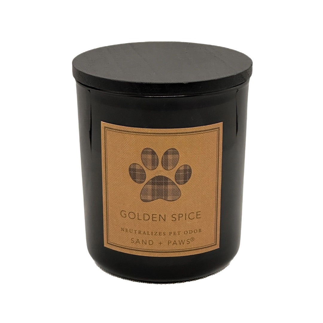 A black glass jar candle with a matching black lid and a tan rectangular label featuring a paw print and the text 'GOLDEN SPICE' and 'SAND + PAWS'.