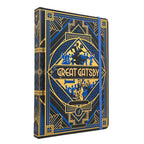 The Great Gatsby hardcover journal featuring an Art Deco design in black, blue, and gold. The cover displays a central diamond-shaped emblem with silhouettes of figures and the title 'The Great Gatsby' in gold lettering.