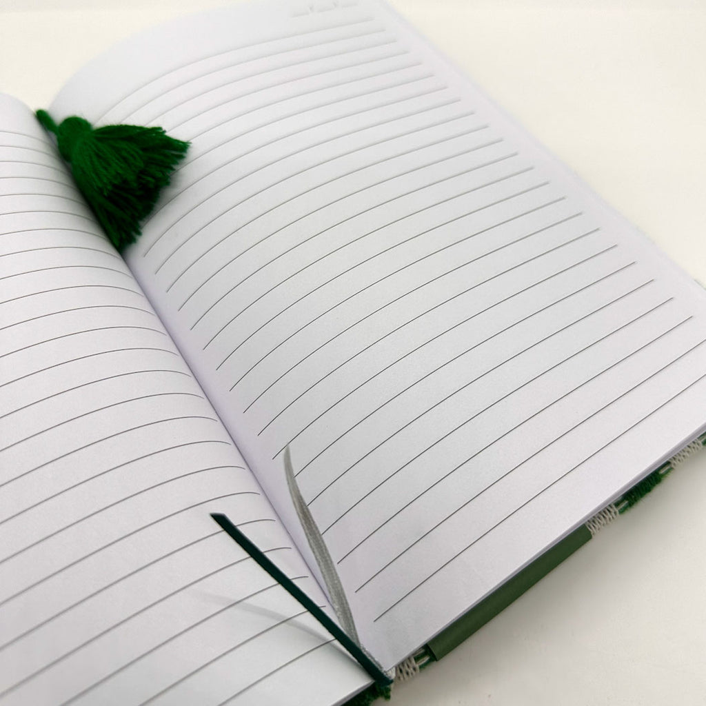 An open lined notebook with white pages, a dark green tassel bookmark, and a green pen loop on a white surface.
