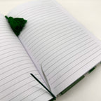 An open lined notebook with white pages, a dark green tassel bookmark, and a green pen loop on a white surface.