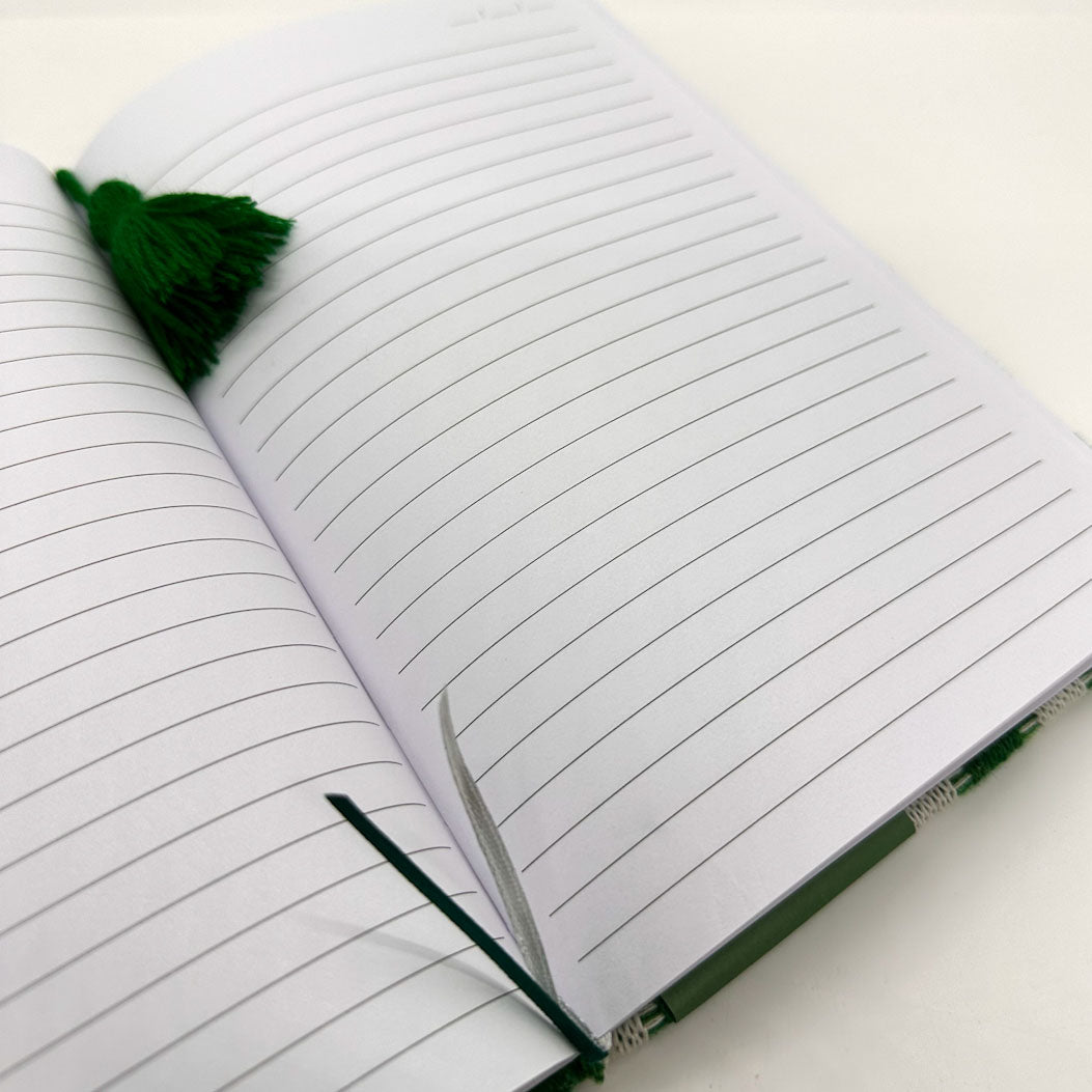 An open lined notebook with white pages, a dark green tassel bookmark, and a green pen loop on a white surface.