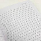 A close-up of a lined notebook page with horizontal black lines on white paper, photographed on a light neutral background.
