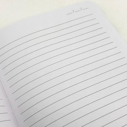 A close-up of a lined notebook page with horizontal black lines on white paper, photographed on a light neutral background.