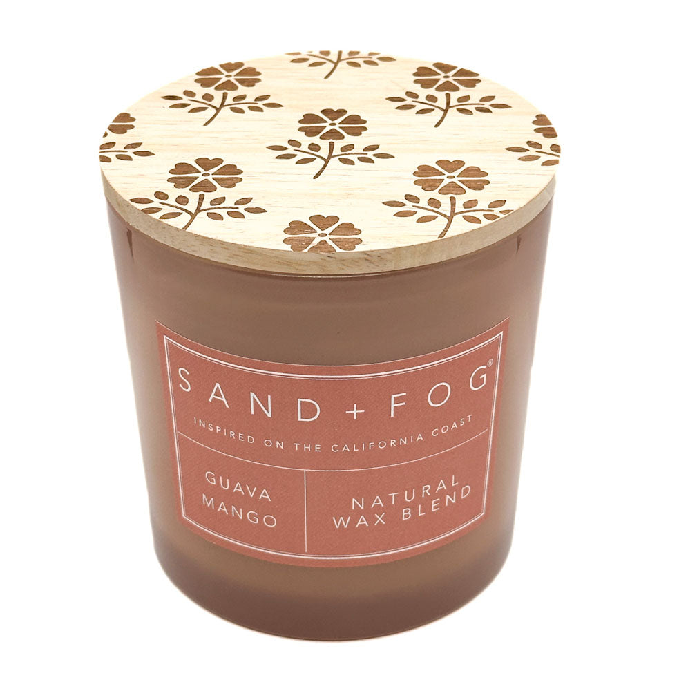 A Sand + Fog Guava Mango natural wax blend candle in a brown glass jar with a light wood lid featuring a brown floral pattern, on a white background.