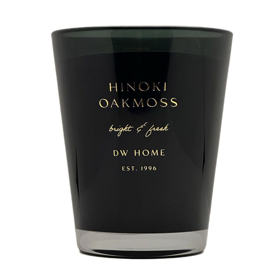 A black glass candle jar with gold lettering that reads 'HINOKI OAKMOSS', 'bright & fresh', and 'DW HOME EST. 1996' on a plain white background.