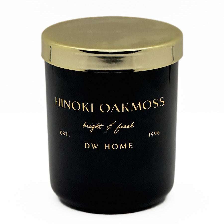 A black glass jar candle with a metallic gold lid, featuring gold lettering that reads 'HINOKI OAKMOSS', 'bright & fresh', 'EST. 1996', and 'DW HOME' on a clean white background.