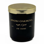 A black glass jar candle with a metallic gold lid, featuring gold lettering that reads 'HINOKI OAKMOSS', 'bright & fresh', 'EST. 1996', and 'DW HOME' on a clean white background.