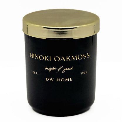 A black glass jar candle with a metallic gold lid, featuring gold lettering that reads 'HINOKI OAKMOSS', 'bright & fresh', 'EST. 1996', and 'DW HOME' on a clean white background.
