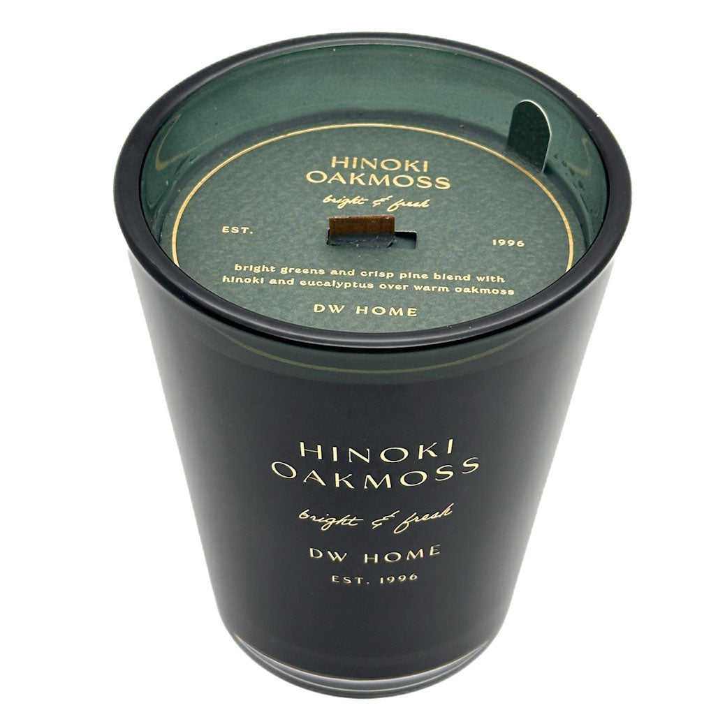 A black glass jar candle with a dark green wax surface and a matching dark green lid. The lid features gold text reading 'HINOKI OAKMOSS bright & fresh' and 'DW HOME'.