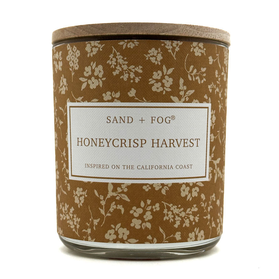 A jar candle with a brown floral-patterned label and a light wood lid, labeled 'SAND + FOG HONEYCRISP HARVEST' on a white background.