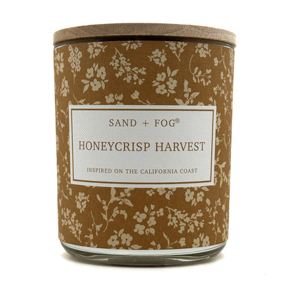 A jar candle with a brown floral-patterned label and a light wood lid, labeled 'SAND + FOG HONEYCRISP HARVEST' on a white background.