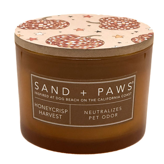 A jar candle from Sand + Paws in the Honeycrisp Harvest scent. The candle features a warm amber-brown translucent glass jar with a matching brown label on the front.
