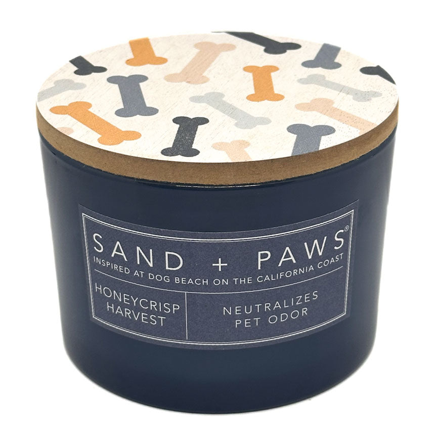A navy blue cylindrical candle jar with a patterned lid featuring dog bones in orange, beige, and grey on a white background. The front label reads 'SAND + PAWS' and 'HONEYCRISP HARVEST'.