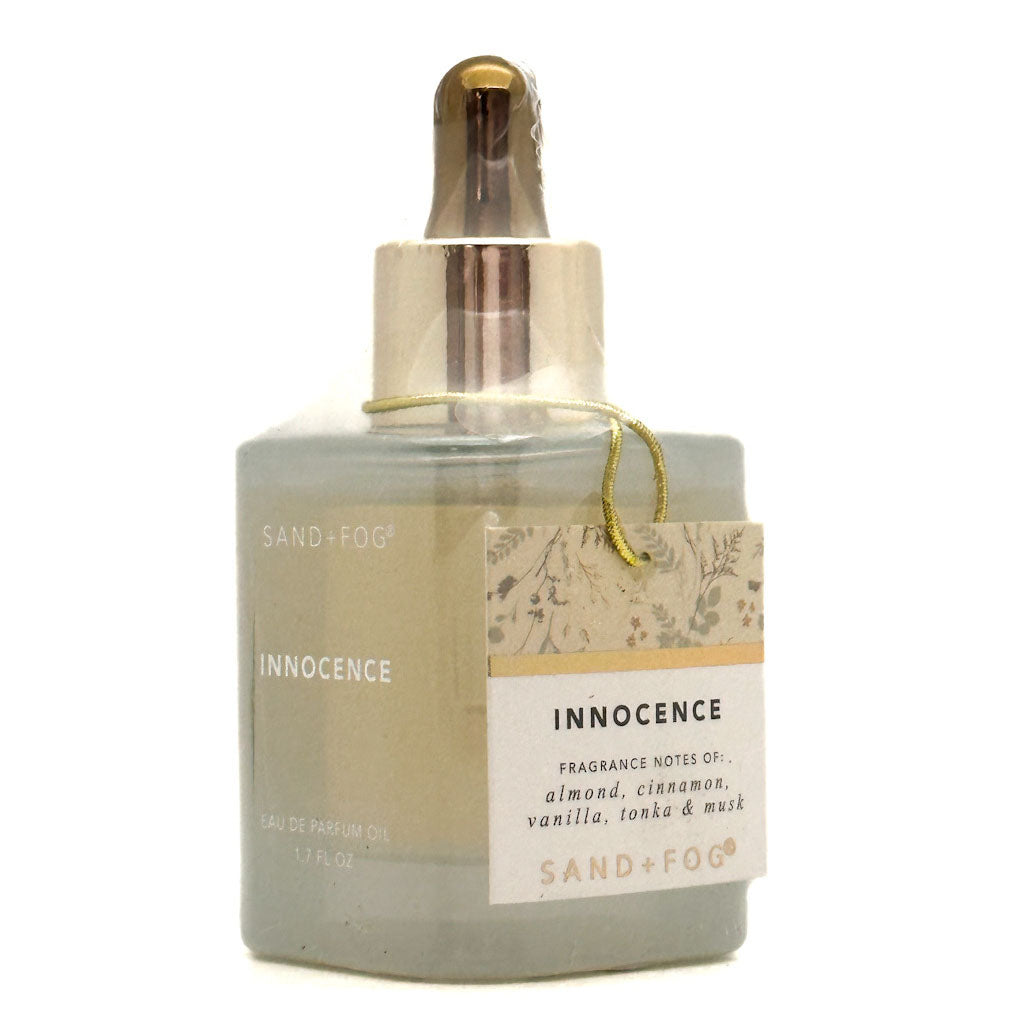 A Sand + Fog Innocence Perfume oil bottle with a dropper cap, featuring a muted sage green-grey glass body, a metallic gold collar, and a cream-coloured paper label with botanical illustrations.