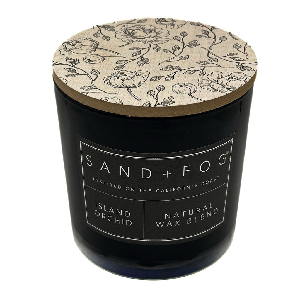 Island Orchid black candle with floral lid labeled 'Sand + Fog' on a white background