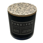 Island Orchid black candle with floral lid labeled 'Sand + Fog' on a white background
