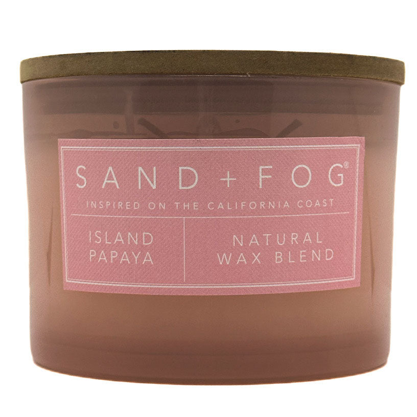 A Sand + Fog candle in a translucent amber-brown glass jar with a muted olive lid and a pink label reading 'ISLAND PAPAYA' and 'NATURAL WAX BLEND'.