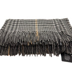 A folded plaid scarf with fringed edges in grey, black, and beige tones, shown on a white background with a small black product tag attached.